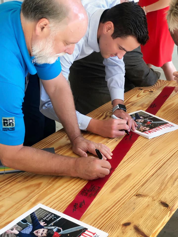 People leaning over wooden table signing a red ribbon with black permanent makers | EnableComp Available Nationwide